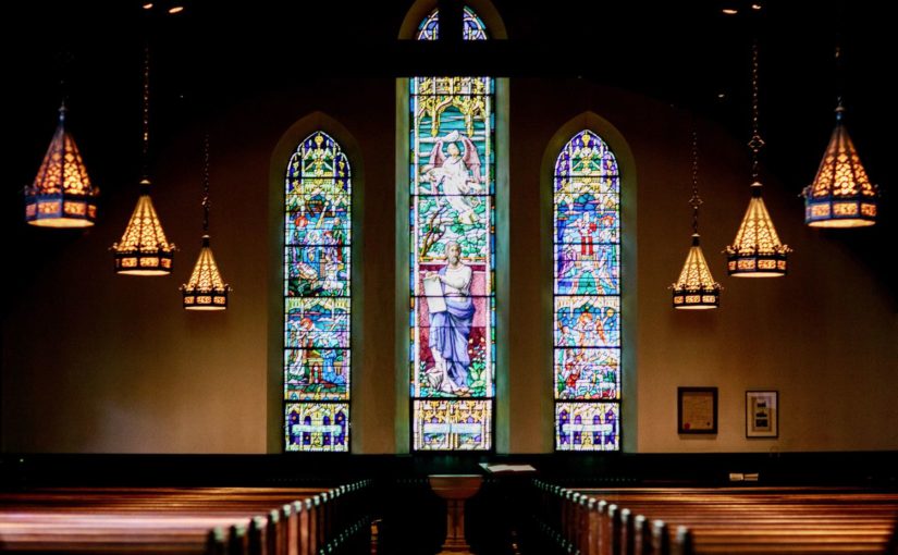 A photograph of the inside of a church featuring stained-glass windows, dark wood pews, and hanging lights.