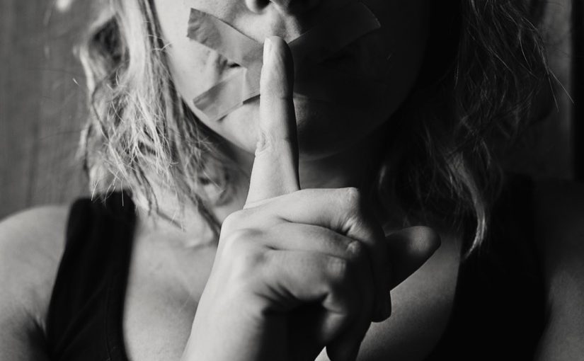 A picture of a woman's face with tape one her lips and a hand signaling, "Shh" ion front of her face. From Nick Byrd's "Sexual Harassment Accusations & The Acceptance Principle"